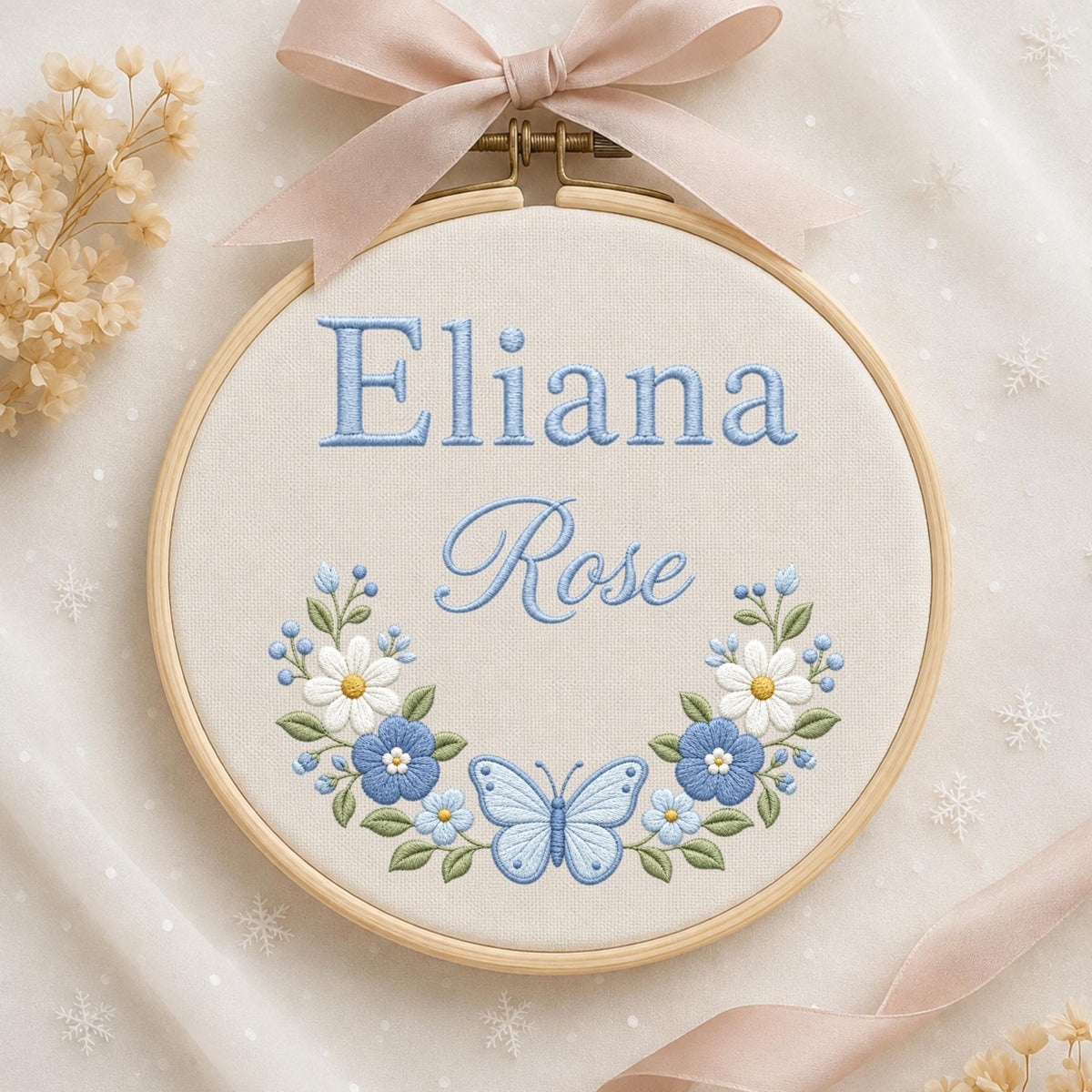 May include: A circular wooden embroidery hoop containing cream-colored fabric with the embroidered blue text Eliana Rose. Below the name is a floral wreath design featuring white and blue flowers, green leaves, and a central light blue butterfly.