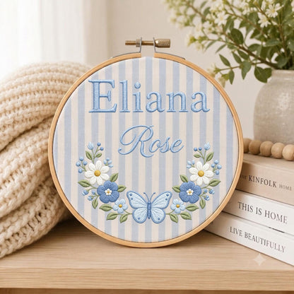 May include: A circular wooden embroidery hoop containing fabric with light blue and white vertical stripes. The text Eliana Rose is embroidered in blue, centered above a floral wreath featuring white and blue flowers and a central blue butterfly.