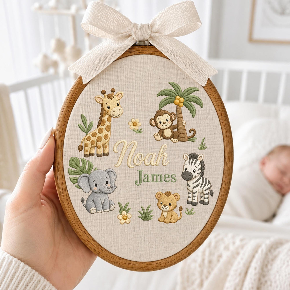 May include: An oval wooden embroidery hoop containing a beige fabric panel with embroidered cartoon animals including a giraffe, elephant, monkey, zebra, and lion. The text Noah James is stitched in the center, topped with a beige fabric bow.