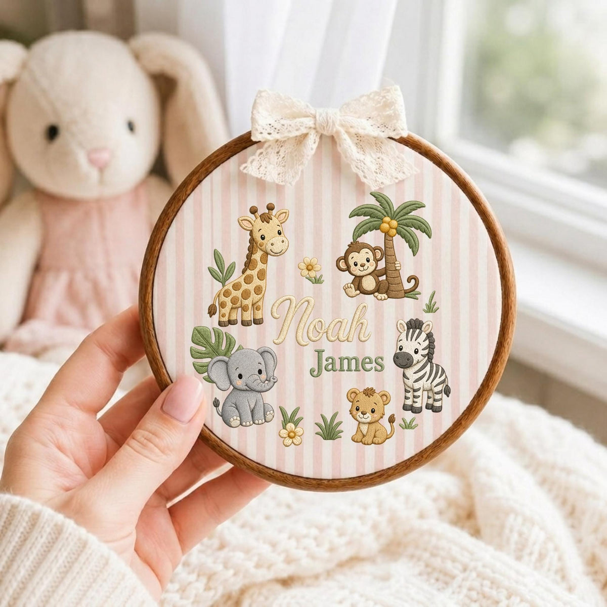 May include: A circular wooden embroidery hoop frame containing fabric with light pink vertical stripes. The fabric features illustrations of a giraffe, monkey, elephant, lion, and zebra, with the text Noah James in the center and a lace bow.