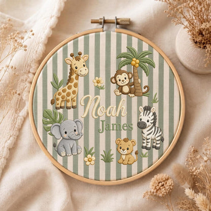 May include: A circular wooden embroidery hoop containing fabric with sage green and white vertical stripes. The fabric features embroidered illustrations of a giraffe, monkey, elephant, zebra, and lion, with the names Noah James in script.