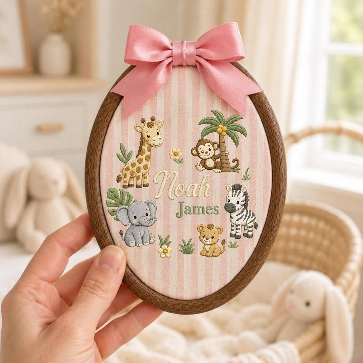 May include: An oval wooden embroidery hoop frame containing fabric with vertical pink and white stripes. The fabric features embroidered illustrations of a giraffe, monkey, elephant, lion, and zebra, with the name Noah James in the center and a pink bow.