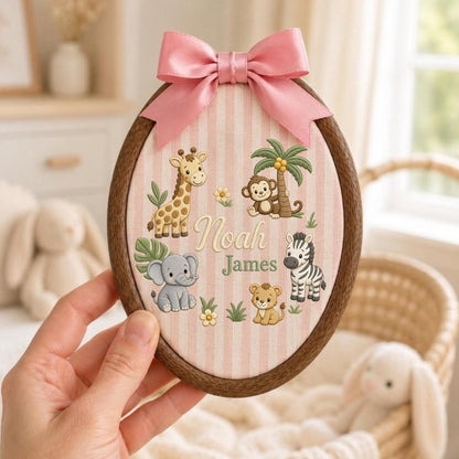May include: An oval wooden embroidery hoop frame containing fabric with vertical pink and white stripes. The fabric features embroidered illustrations of a giraffe, monkey, elephant, lion, and zebra, with the name Noah James in the center and a pink bow.
