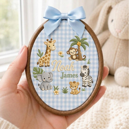May include: An oval wooden embroidery hoop featuring a light blue and white gingham fabric background. The fabric is embroidered with a giraffe, monkey, elephant, zebra, and lion, along with the names Noah James in script and block lettering.