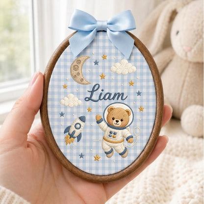 May include: An oval wooden embroidery hoop featuring a light blue and white gingham fabric background. The design includes an embroidered astronaut bear, a rocket ship, a crescent moon, clouds, and stars. The name Liam is stitched in blue.