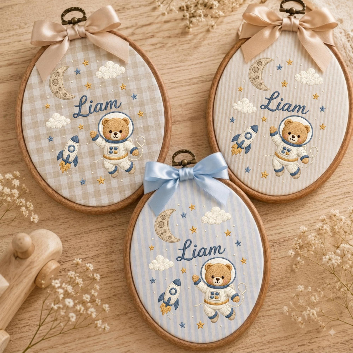 May include: Three oval embroidery hoops featuring a brown bear astronaut, a rocket ship, a crescent moon, clouds, and stars. Each hoop is personalized with the name Liam in blue script and topped with a decorative satin ribbon bow.