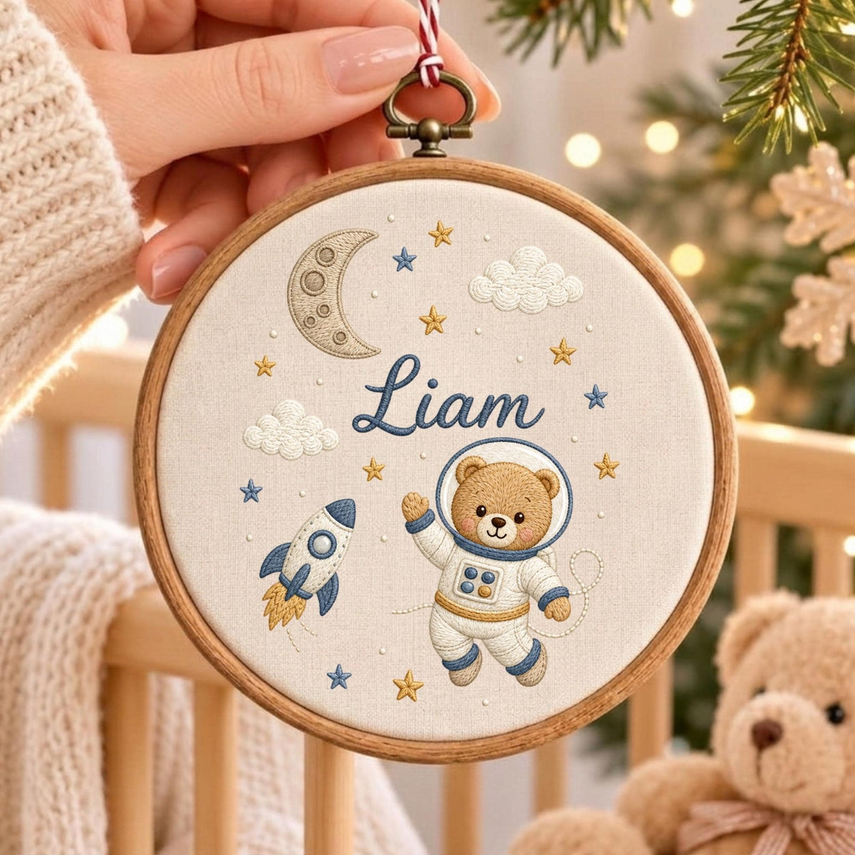 May include: A round wooden embroidery hoop containing cream-colored fabric with an embroidered space theme. The design features a brown teddy bear in a white astronaut suit, a blue and yellow rocket, a crescent moon, clouds, and stars with the name Liam.