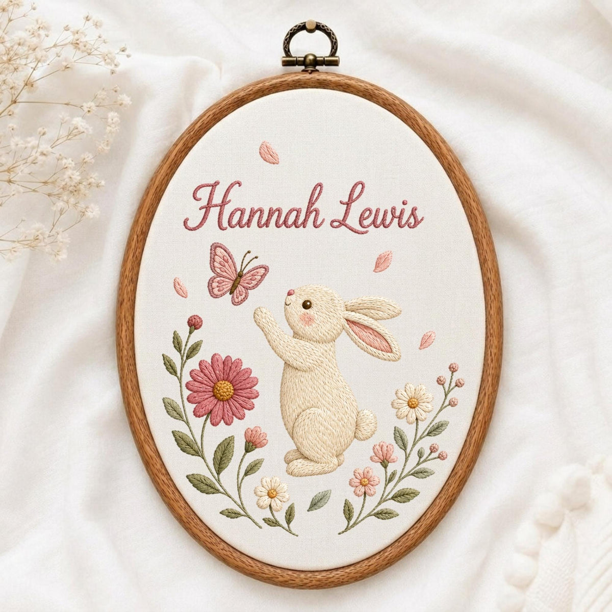 May include: An oval wooden embroidery hoop containing white fabric with a stitched design of a cream-colored rabbit reaching for a pink butterfly. The name Hannah Lewis is embroidered in pink script above the rabbit, surrounded by pink and white flowers.
