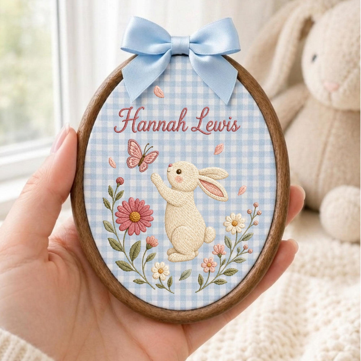 May include: An oval wooden embroidery hoop featuring a light blue and white gingham fabric background. The embroidery depicts a cream-colored rabbit reaching for a pink butterfly, surrounded by pink and white flowers. The name Hannah Lewis is stitched in pink.