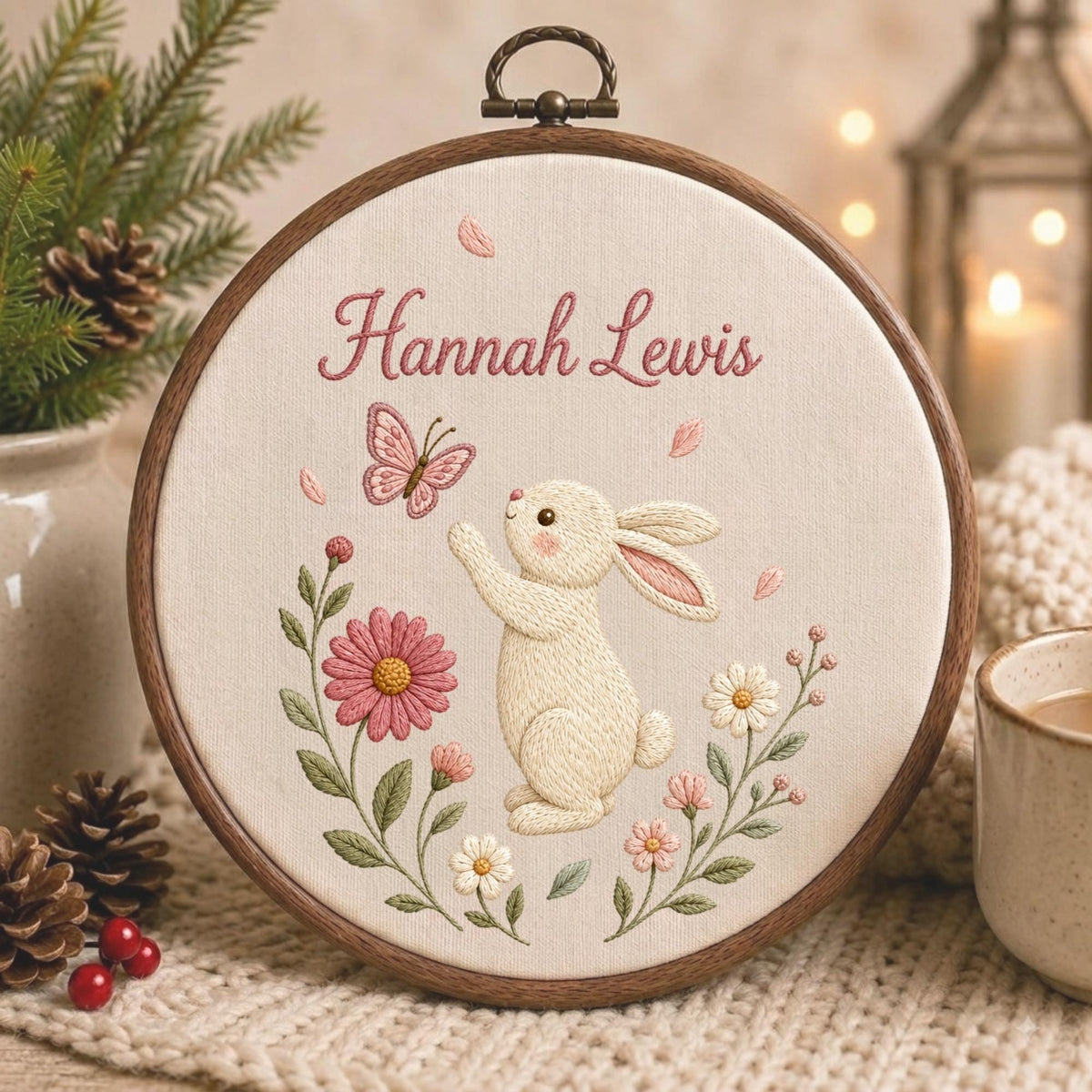 May include: A circular wooden embroidery hoop containing cream-colored fabric with a stitched design of a white rabbit reaching toward a pink butterfly. The text Hannah Lewis is embroidered in pink above the rabbit, surrounded by pink and white flowers.