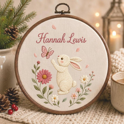 May include: A circular wooden embroidery hoop containing cream-colored fabric with a stitched design of a white rabbit reaching toward a pink butterfly. The text Hannah Lewis is embroidered in pink above the rabbit, surrounded by pink and white flowers.