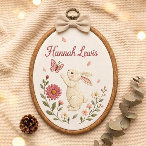 May include: An oval wooden embroidery hoop containing white fabric with a stitched design of a cream-colored rabbit reaching for a pink butterfly. The name Hannah Lewis is embroidered in pink script above the rabbit, surrounded by pink and white flowers.