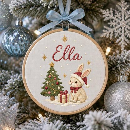 May include: A circular wooden embroidery hoop containing a cream-colored fabric with the name Ella embroidered in red script. The design features a green Christmas tree with ornaments, a gift box, and a beige rabbit wearing a red Santa hat.