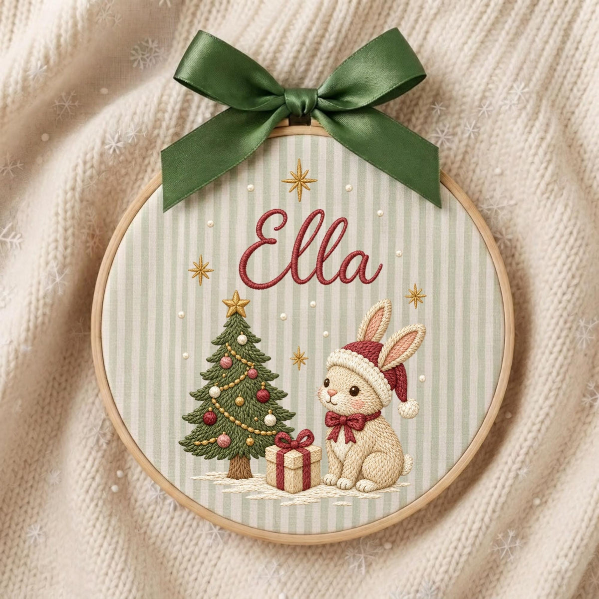 May include: A round wooden embroidery hoop featuring a light green and white striped fabric background. The embroidery displays the name Ella in red script, a decorated Christmas tree, a gift box, and a rabbit wearing a red Santa hat and bow.
