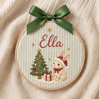 May include: A round wooden embroidery hoop featuring a light green and white striped fabric background. The embroidery displays the name Ella in red script, a decorated Christmas tree, a gift box, and a rabbit wearing a red Santa hat and bow.