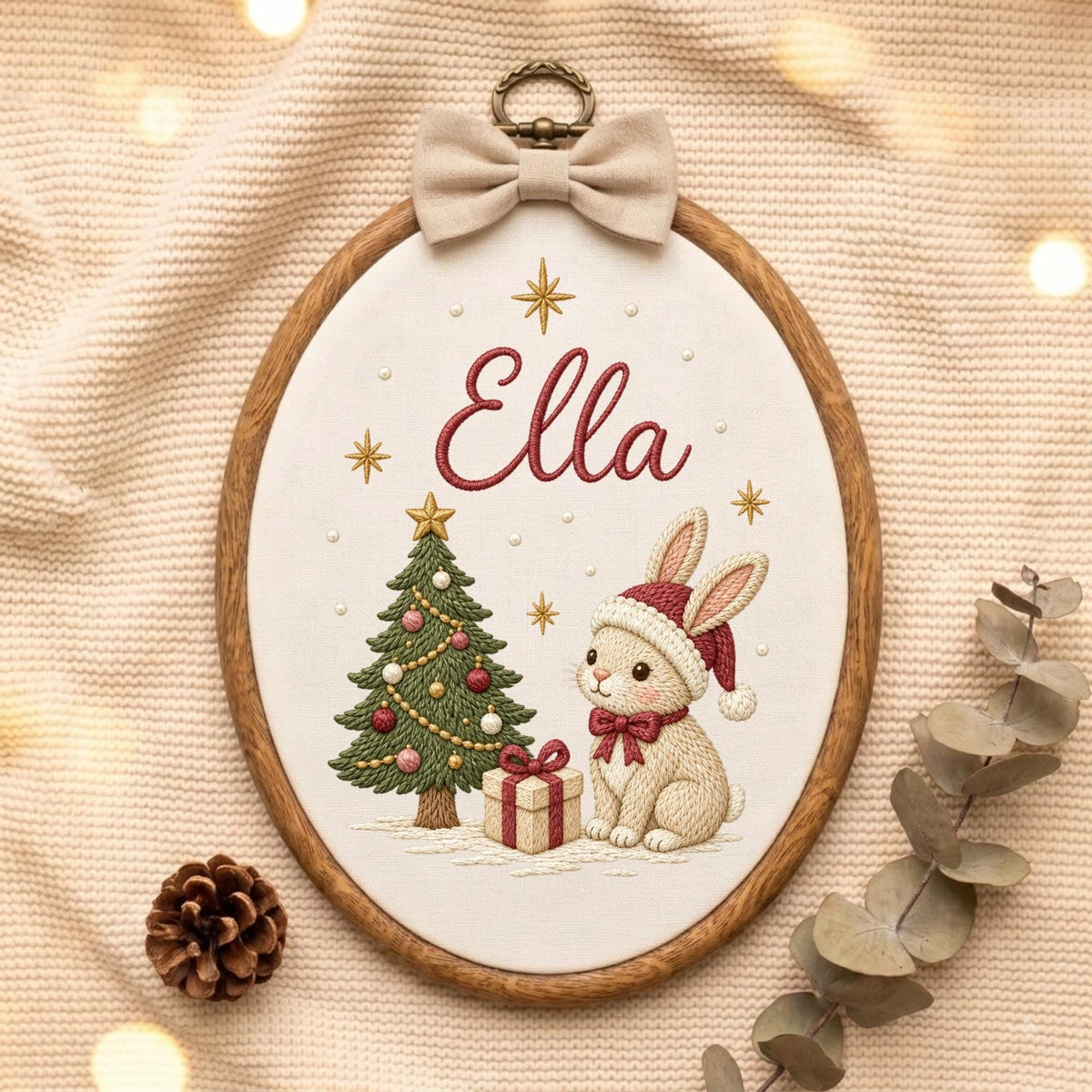 May include: An oval wooden embroidery hoop containing white fabric with the embroidered name Ella in red script. The design features a green Christmas tree with ornaments, a rabbit wearing a red Santa hat and bow, and a gift box with a red ribbon.