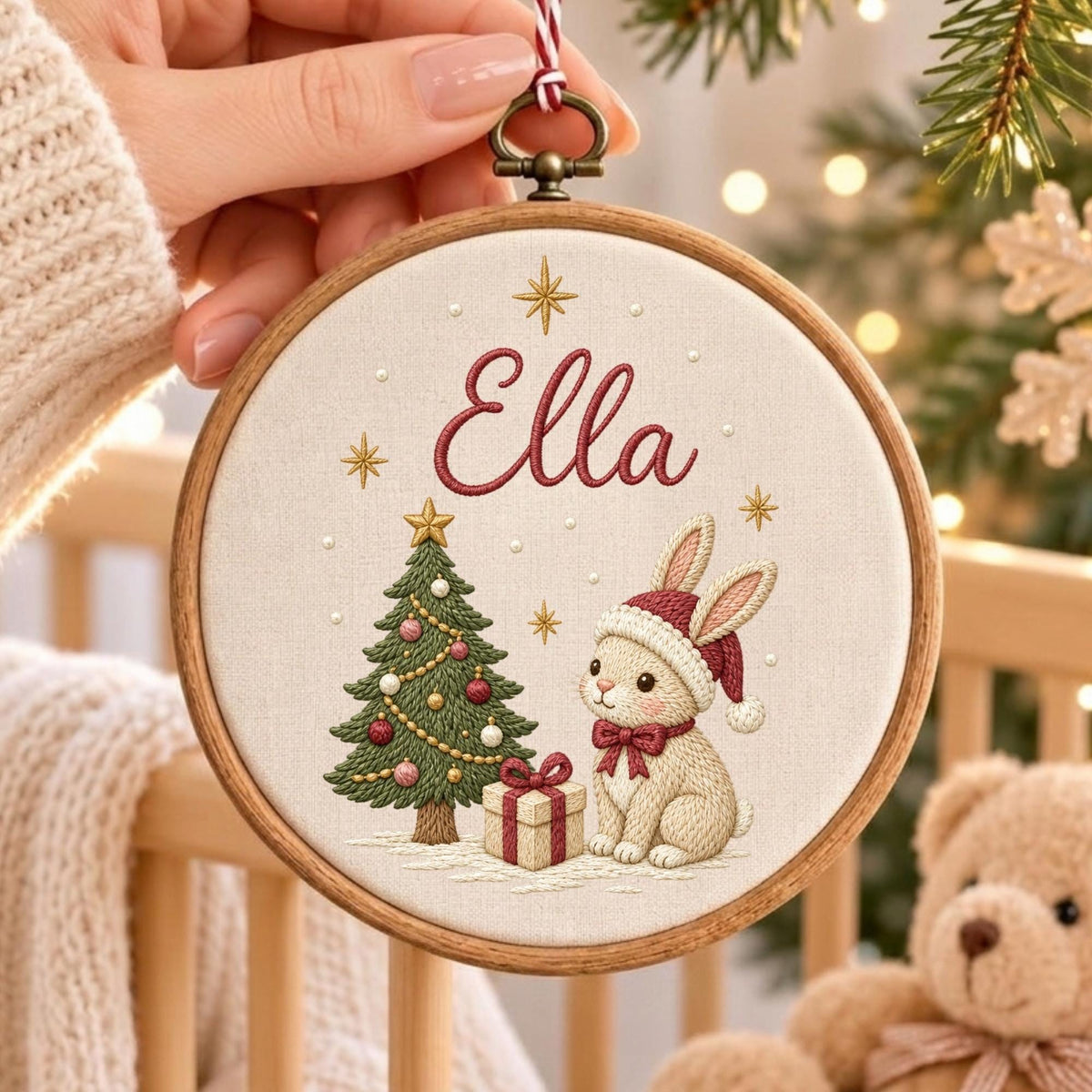 May include: A circular wooden embroidery hoop containing a cream-colored fabric panel. The fabric features an embroidered Christmas tree, a gift box, and a rabbit wearing a red hat. The name Ella is stitched in red script above the design.