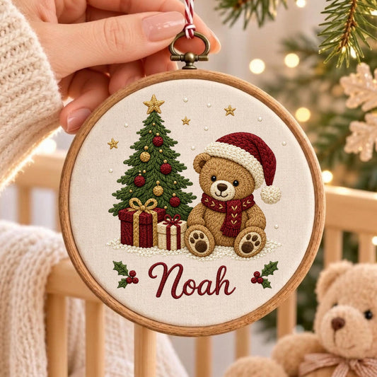 May include: A round wooden embroidery hoop featuring a cream-colored fabric with a stitched design of a brown teddy bear wearing a red Santa hat and scarf, a decorated Christmas tree, two gift boxes, and the name Noah in red cursive script.