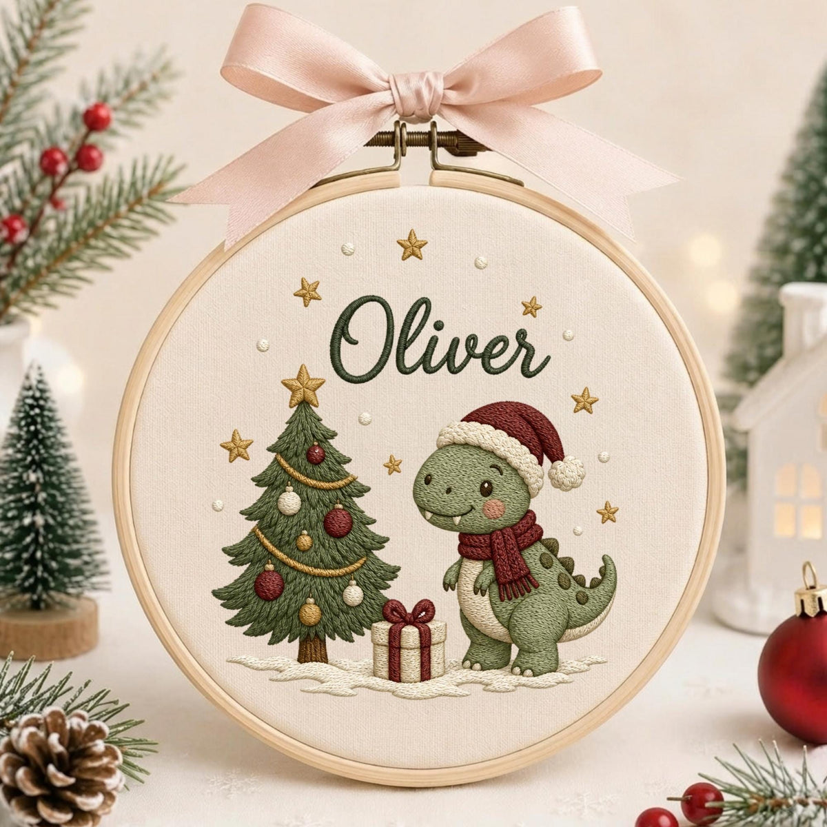 May include: A round wooden embroidery hoop featuring a cream fabric background with a stitched green dinosaur wearing a red Santa hat and scarf, a decorated Christmas tree, a gift box, and the name Oliver, topped with a pale pink ribbon bow.