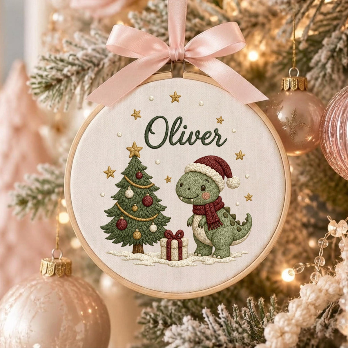 May include: A circular embroidery hoop ornament featuring a light-colored fabric background with the name Oliver embroidered in dark green. The design includes a green Christmas tree, a green dinosaur wearing a red hat and scarf, and a gift box.