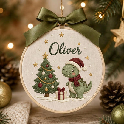 May include: A round wooden embroidery hoop ornament featuring a cream-colored fabric background. The design shows a green dinosaur wearing a red Santa hat and scarf, a decorated Christmas tree, a gift box, and the name Oliver in green text.
