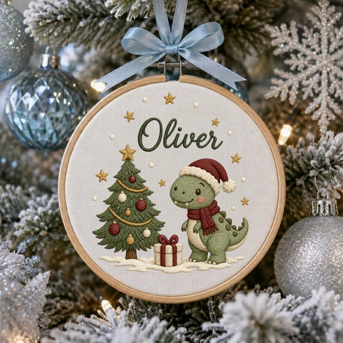 May include: A round wooden embroidery hoop ornament featuring a light fabric background with the embroidered name Oliver, a green Christmas tree with red and gold ornaments, a gift box, and a green dinosaur wearing a red Santa hat and scarf.