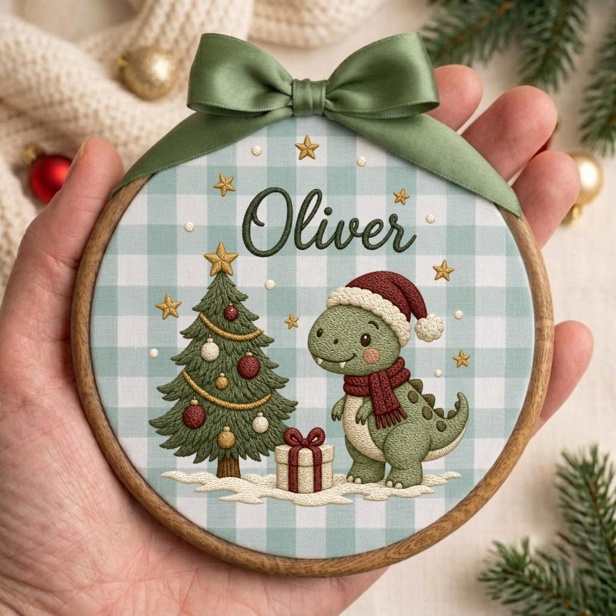 May include: A circular wooden embroidery hoop ornament featuring a light blue and white gingham fabric background. The design includes a green dinosaur wearing a red Santa hat and scarf, a decorated Christmas tree, and the name Oliver in script.