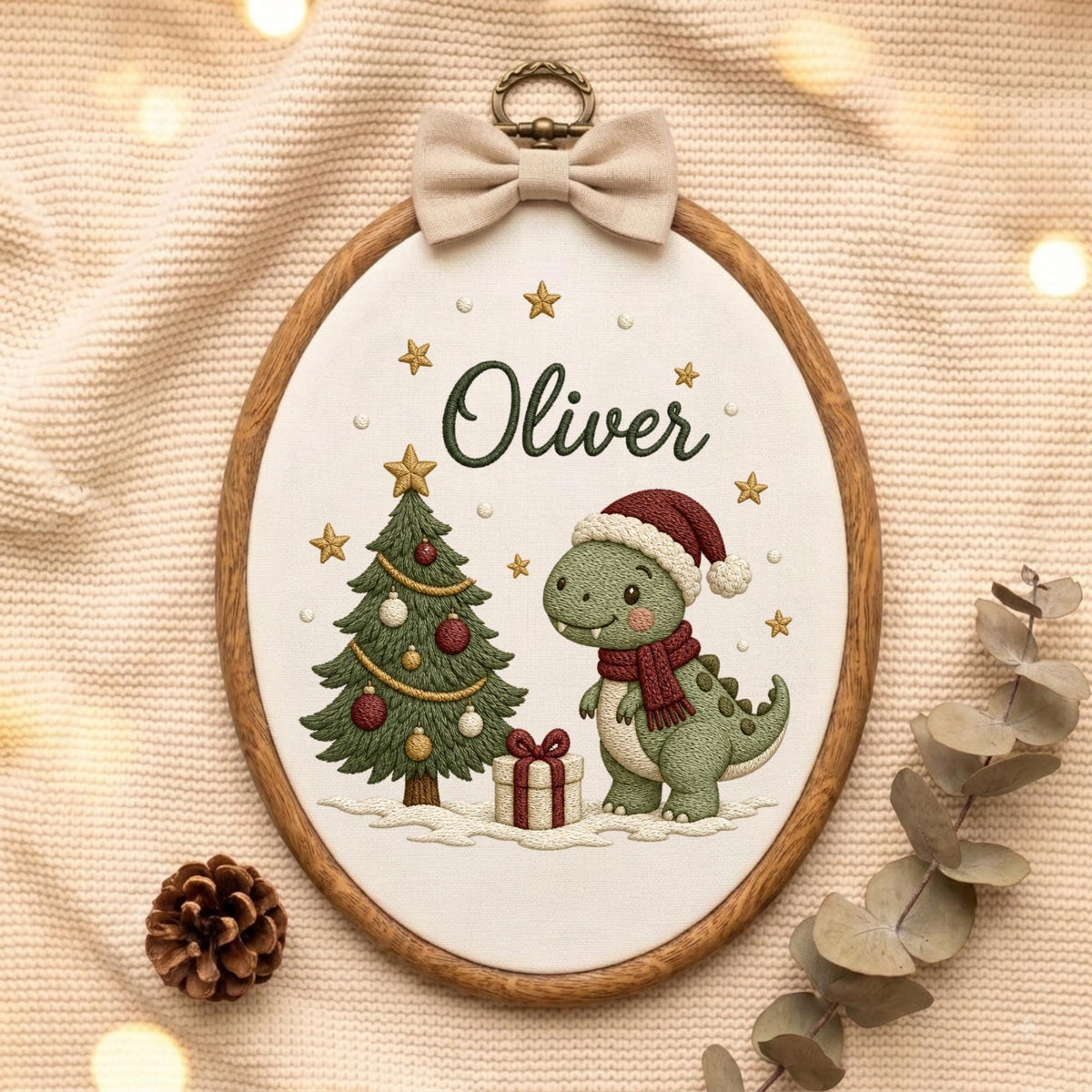 May include: An oval embroidery hoop featuring a green dinosaur wearing a red Santa hat and scarf, standing next to a decorated Christmas tree and a gift box. The name Oliver is stitched in dark green above the dinosaur, with gold stars.