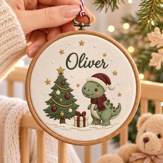 May include: A round wooden embroidery hoop containing a cream-colored fabric with a stitched design of a green dinosaur wearing a red Santa hat and scarf, a decorated Christmas tree, a gift box, and the name Oliver in dark green script.