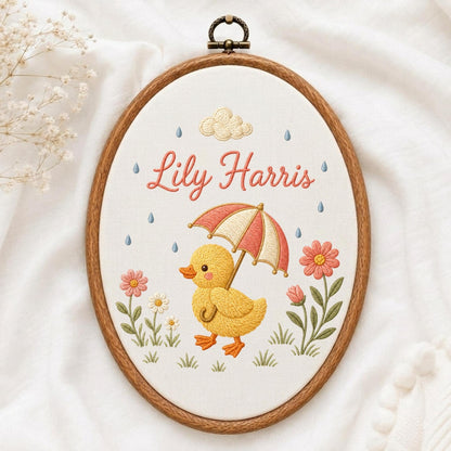 May include: An oval embroidery hoop containing white fabric with a stitched design of a yellow duckling holding a red and white striped umbrella. The text Lily Harris is embroidered in pink above the duckling, surrounded by flowers and raindrops.
