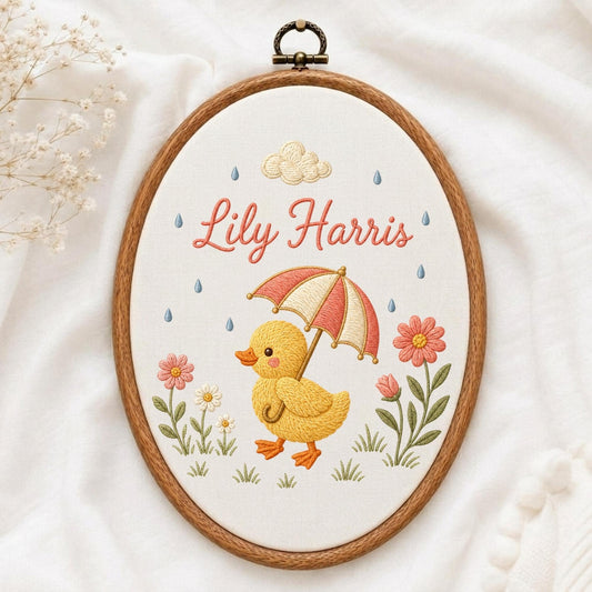 May include: An oval embroidery hoop containing white fabric with a stitched design of a yellow duckling holding a red and white striped umbrella. The text Lily Harris is embroidered in pink above the duckling, surrounded by flowers and raindrops.