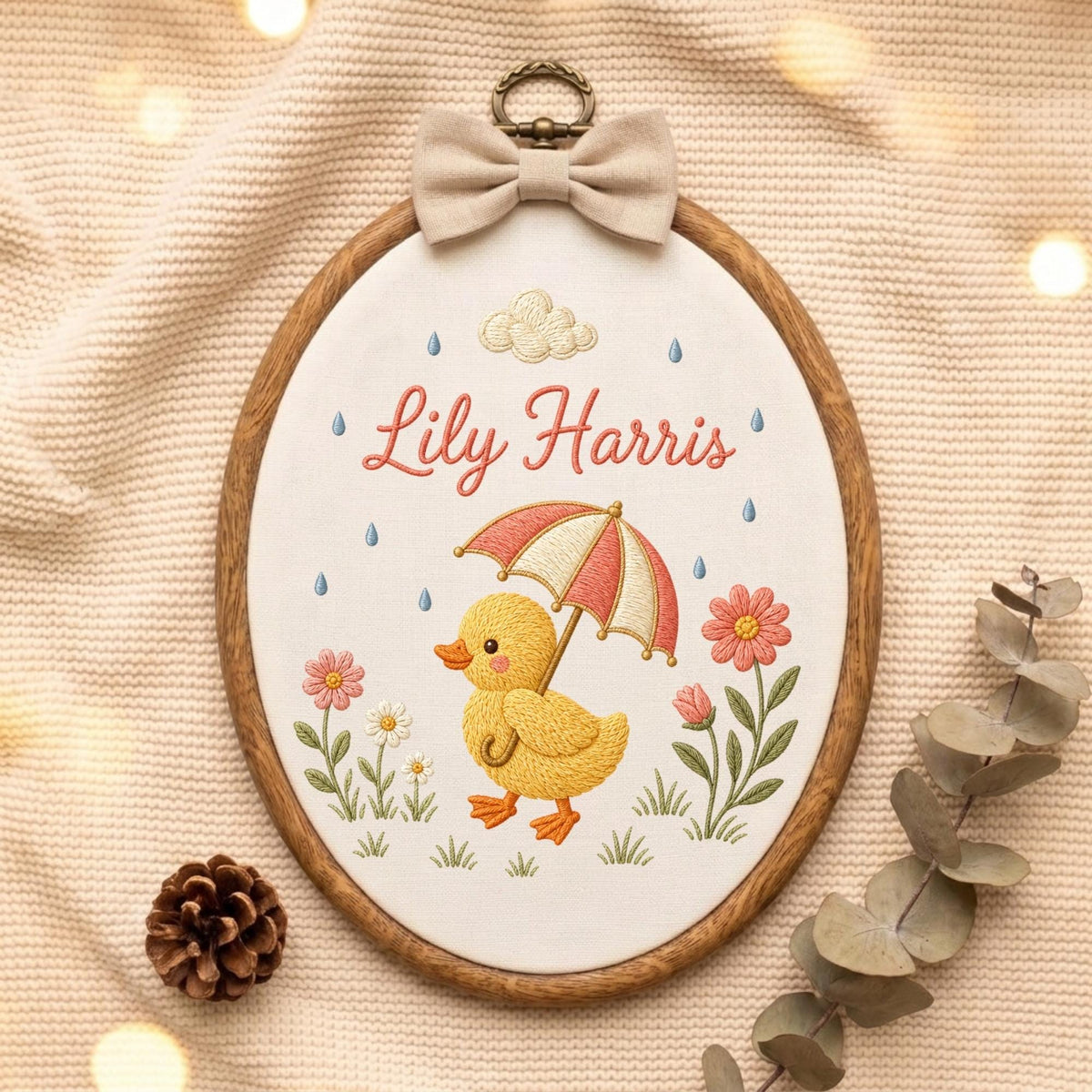 May include: An oval wooden embroidery hoop containing white fabric with a stitched design of a yellow duckling holding a red and white striped umbrella. The text Lily Harris is embroidered in red above the duckling, surrounded by flowers and rain.