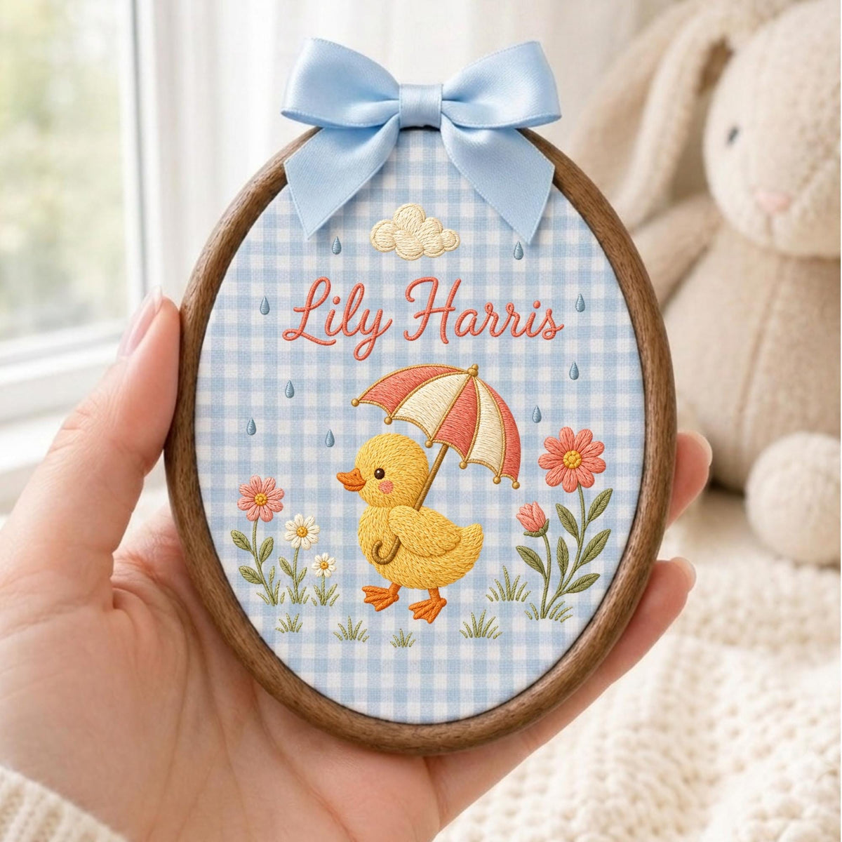 May include: An oval embroidery hoop featuring a light blue and white gingham fabric background. The design includes a yellow duckling holding a red and white umbrella, pink and white flowers, a cloud, and the embroidered name Lily Harris.