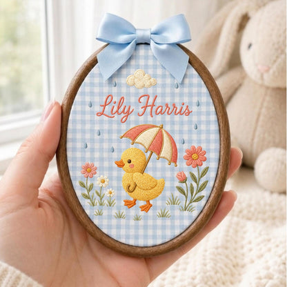 May include: An oval embroidery hoop featuring a light blue and white gingham fabric background. The design includes a yellow duckling holding a red and white umbrella, pink and white flowers, a cloud, and the embroidered name Lily Harris.