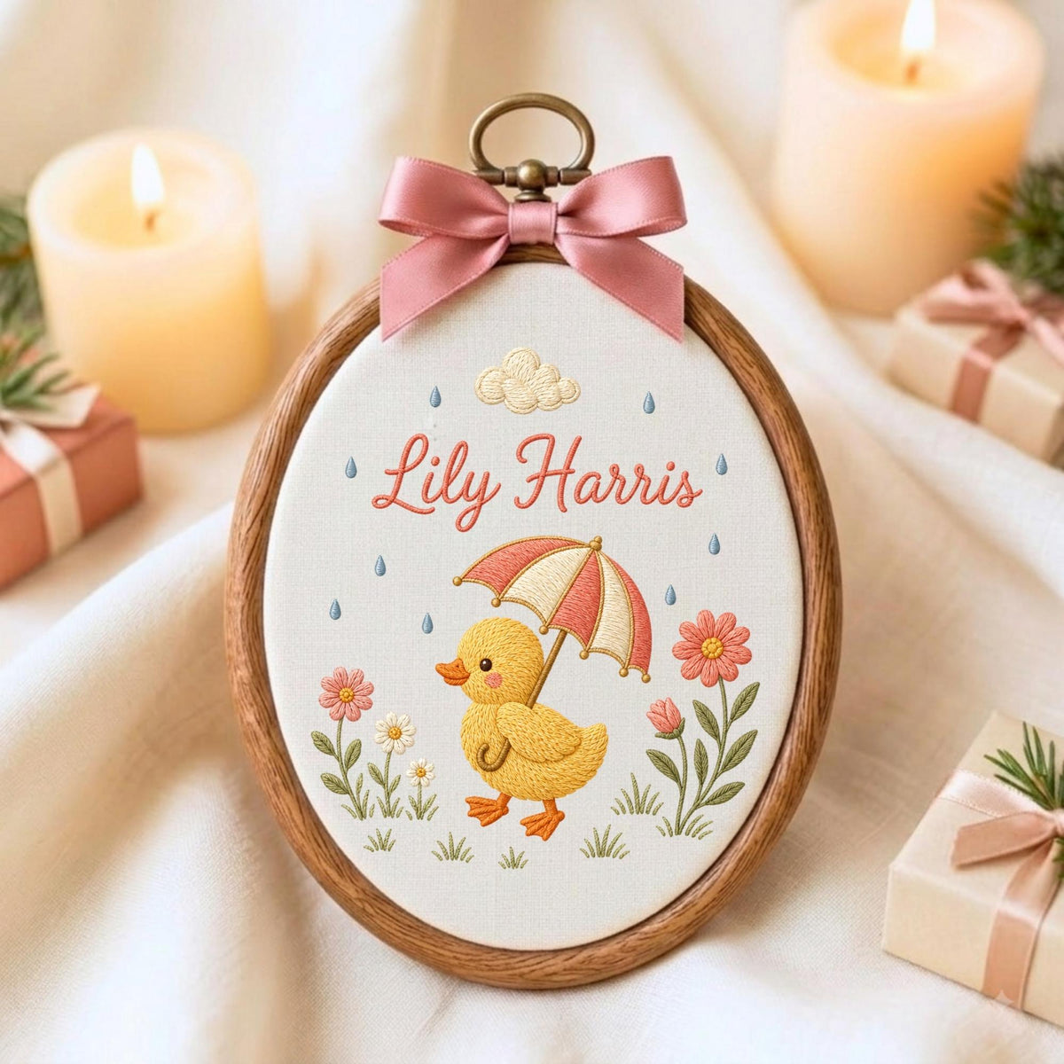 May include: An oval wooden embroidery hoop containing white fabric with a hand-stitched design of a yellow duckling holding a red and white striped umbrella. The name Lily Harris is embroidered in red script above the duckling, with flowers.