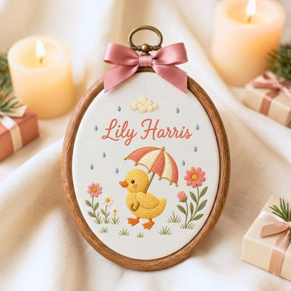 May include: An oval wooden embroidery hoop containing white fabric with a hand-stitched design of a yellow duckling holding a red and white striped umbrella. The name Lily Harris is embroidered in red script above the duckling, with flowers.