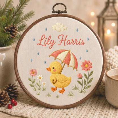 May include: A circular wooden embroidery hoop containing a cream-colored fabric with a stitched design of a yellow duckling holding a red and white striped umbrella. The text Lily Harris is embroidered in pink above the duckling, with flowers and raindrops.
