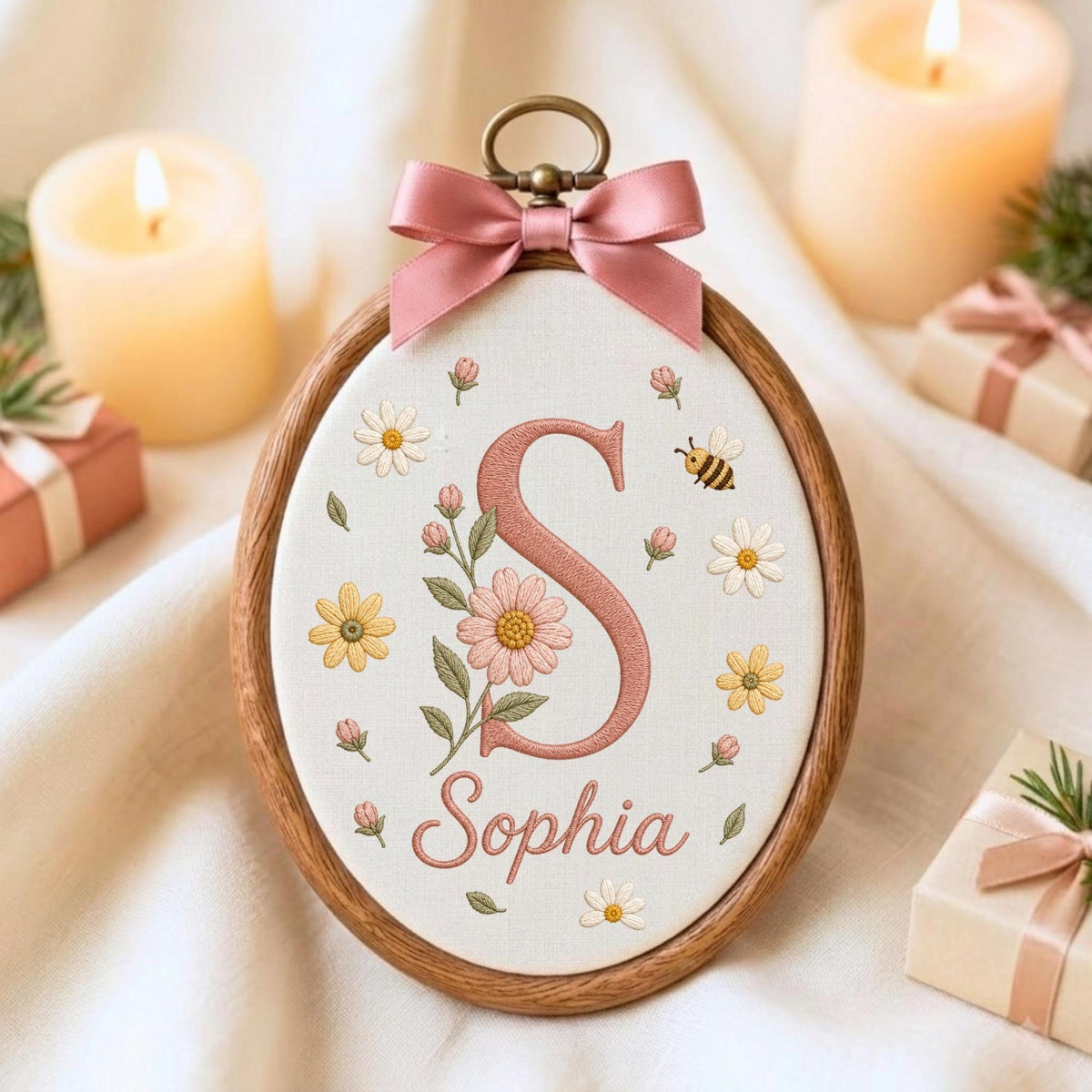 May include: An oval wooden embroidery hoop containing white fabric with a stitched pink letter S, the name Sophia, and floral motifs including daisies, buds, and a small bee. A dusty pink satin ribbon bow is attached to the top metal hanger.