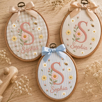 May include: Three oval wooden embroidery hoops featuring fabric backgrounds with embroidered pink letters S, the name Sophia, daisies, and bees. The hoops are topped with satin bows in beige, light blue, and cream, displaying floral motifs.