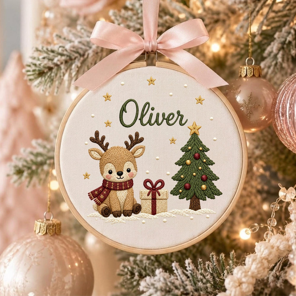 May include: A circular embroidery hoop ornament featuring a light brown reindeer wearing a red scarf, a green Christmas tree with red and gold ornaments, a gift box with a red bow, and the name Oliver in green text, topped with a pink ribbon.