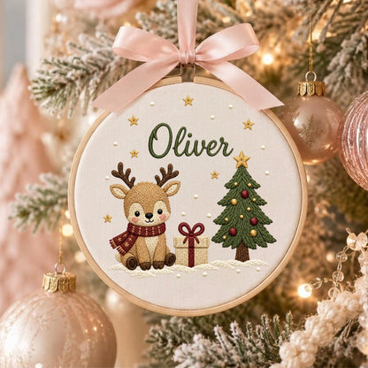 May include: A circular embroidery hoop ornament featuring a light brown reindeer wearing a red scarf, a green Christmas tree with red and gold ornaments, a gift box with a red bow, and the name Oliver in green text, topped with a pink ribbon.