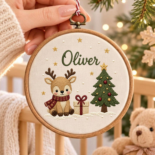 May include: A circular wooden embroidery hoop containing a cream-colored fabric panel. The embroidery features a brown reindeer wearing a red scarf, a gift box with a red ribbon, and a green Christmas tree. The name Oliver is stitched in green.