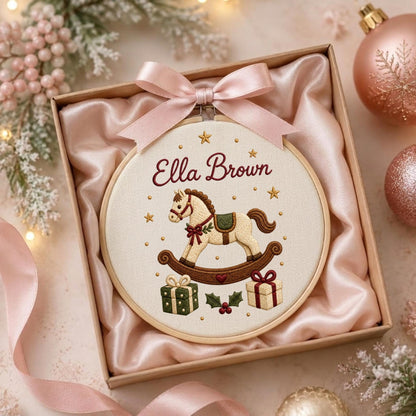 May include: A round wooden embroidery hoop containing a cream-colored fabric with the embroidered name Ella Brown, a rocking horse, two gift boxes, and small stars. A light pink satin ribbon is tied in a bow at the top of the hoop.