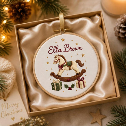 May include: A round wooden embroidery hoop containing white fabric with a stitched rocking horse, the name Ella Brown, and Christmas gifts. The hoop is displayed inside a square box lined with champagne-colored satin fabric.
