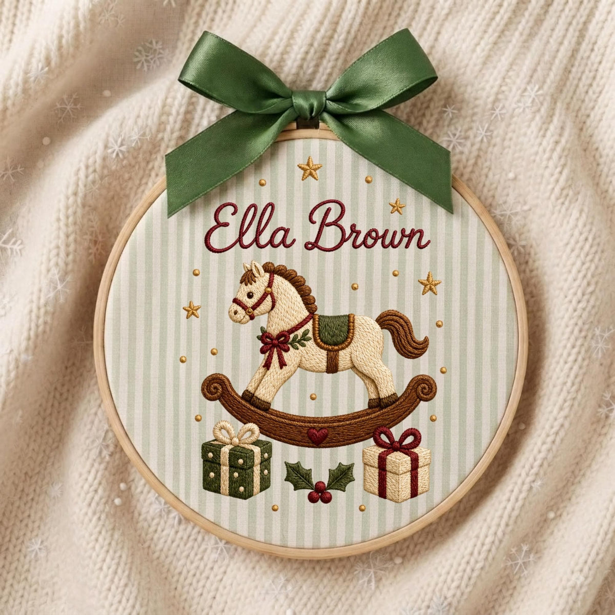 May include: A circular embroidery hoop featuring a light green and white striped fabric background. The design includes the embroidered name Ella Brown, a rocking horse, two gift boxes, holly leaves, and small gold stars, topped with a green ribbon.