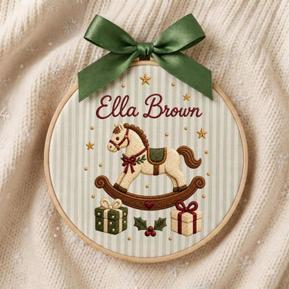 May include: A circular embroidery hoop featuring a light green and white striped fabric background. The design includes the embroidered name Ella Brown, a rocking horse, two gift boxes, holly leaves, and small gold stars, topped with a green ribbon.