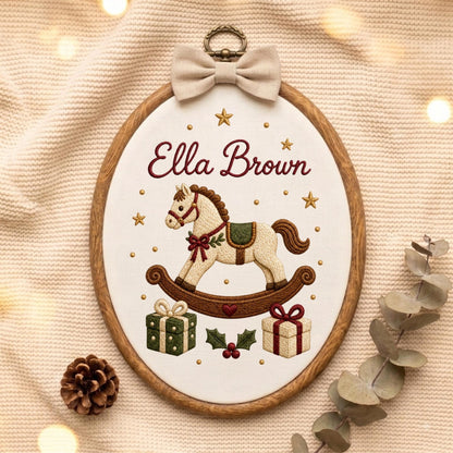 May include: An oval wooden embroidery hoop containing white fabric with the embroidered name Ella Brown in dark red script. The design features a cream-colored rocking horse, two gift boxes, holly leaves, and gold stars, topped with a beige bow.