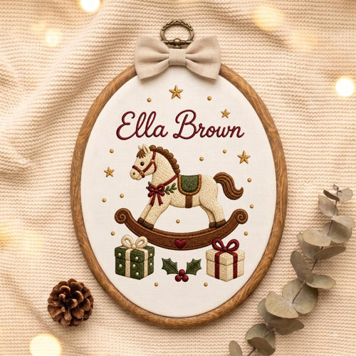 May include: An oval wooden embroidery hoop containing white fabric with the embroidered name Ella Brown in dark red script. The design features a cream-colored rocking horse, two gift boxes, holly leaves, and gold stars, topped with a beige bow.