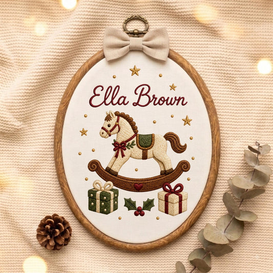 May include: An oval wooden embroidery hoop containing white fabric with the embroidered name Ella Brown in dark red script. The design features a cream-colored rocking horse, two gift boxes, holly leaves, and gold stars, topped with a beige bow.
