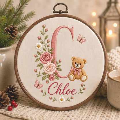 May include: A circular wooden embroidery hoop containing cream-colored fabric with a stitched design. The design features a large pink letter C, a brown teddy bear with a pink bow, pink roses, white daisies, and two small pink butterflies.
